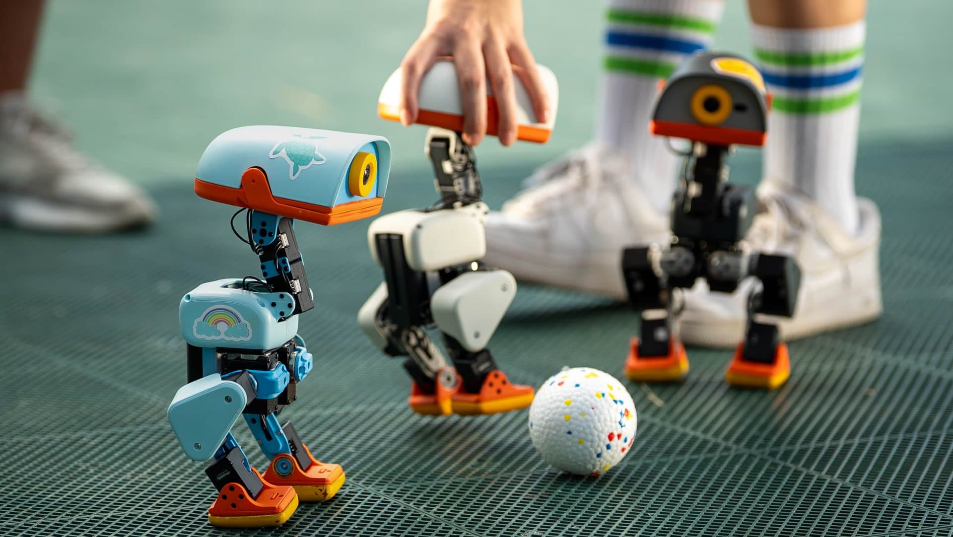 Three small bipedal Microduck robots around a ball on a green sports floor