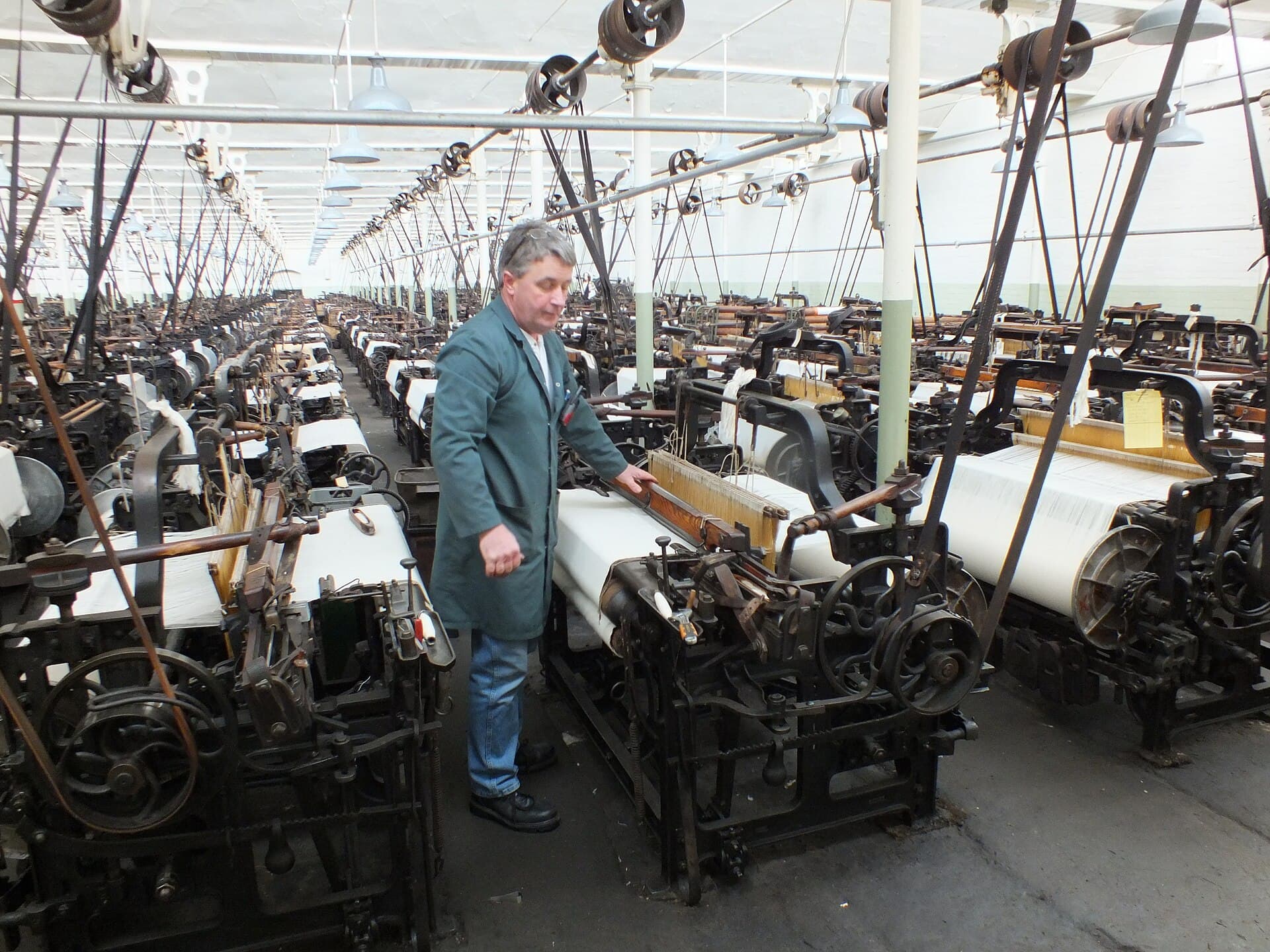 A long weaving shed of belt-driven power looms, with one weaver at a loom in the foreground